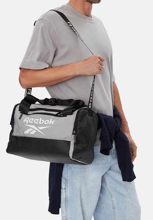 Hombre con camiseta gris claro y vaqueros azul claro lleva un bolso de lona Reebok gris y negro sobre el hombro.