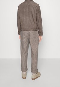 Ruskea nubukkinahkakangas takki kauluksella, yhdistettynä beigen ja ruskean ruudullisiin housuihin sekä vaalean beigen värisiin liukuvaan kenkään.