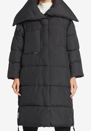 Zwarte lange pufferjas met grote kraag, voorzak op de borst, lange mouwen en ritssluitingen in de zijnaden, gedragen over een witte geribbelde coltrui.