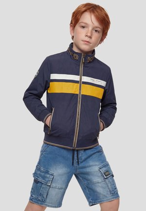 Niño pelirrojo con chaqueta azul marino con rayas amarillas y blancas y pantalones cortos de mezclilla tipo cargo, de pie con las manos en los bolsillos.