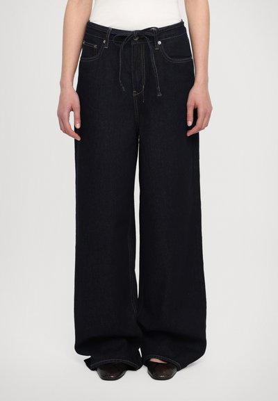 Jeans en denim foncé à jambes larges avec une taille à cordon, dotés de poches avant et de coutures contrastées le long des coutures. Tissu texturé.