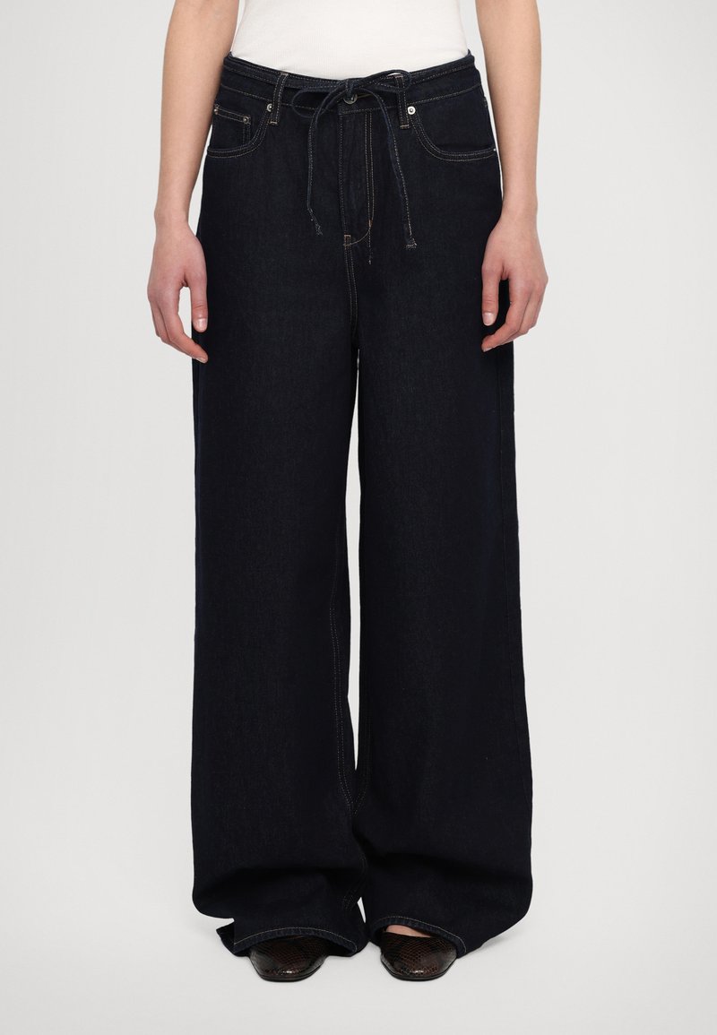 Vide benbukser i mørkt denim med snor i livet, med frontlommer og kontraststing langs sømmene. Teksturert stoff.