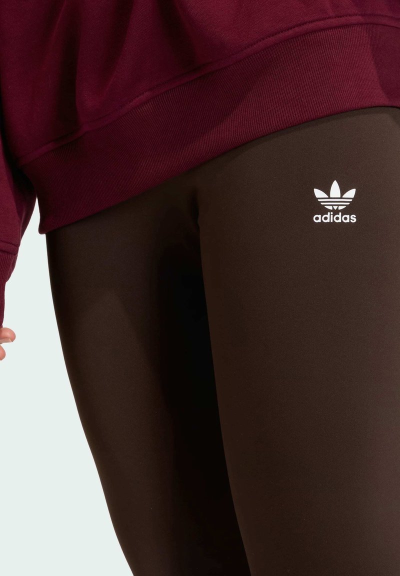 Leggings marroni con una texture liscia e vestibilità elastica, con un logo bianco Adidas sul lato e abbinati a una felpa bordeaux.