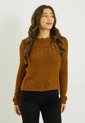 Joven con cabello largo y ondulado, vistiendo un suéter de punto con patrón color óxido y pantalones negros, mirando de lado sobre un fondo liso.