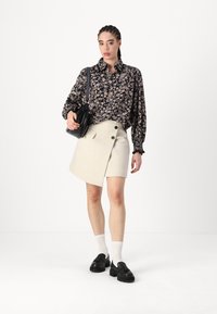 Blusa negra con estampado floral y mangas largas, combinada con una falda envolvente beige que presenta dos botones oscuros. Los accesorios incluyen un bolso negro y zapatos.