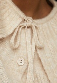 Pull crème avec une texture tricotée, fermeture à boutons et col plat. Dispose d'un nœud au cou et d'un tissu doux.