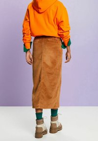 Brauner Cord-Midi-Rock mit vertikalen Rippungen, kombiniert mit einem orangefarbenen Hoodie und beigen Schnürstiefeln. Hintergrund in sanften lila Farbtönen.