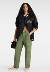 Schwarze Bomberjacke mit weißen Streifen und Logo, kombiniert mit grünen Cargo-Hosen mit Seitentaschen und rosa karierter Slip-On-Schuhe.