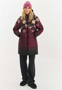 Prošívaný kabát s květinovým potiskem ve tmavé burgundské a černé barvě, s předním zipem, kapucí a dvěma kapsami. Modelka má na sobě černé džíny a tmavé boty.