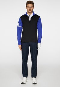 Svart och blå tröja med zip-krage, lång ärm och randiga detaljer på vänster ärm; ihop med marinblå cargobyxor och vita skor.