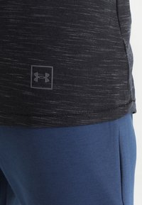 Svart kortärmad skjorta med ett strukturerat mönster och en Under Armour-logga, kombinerat med blåa sweats.