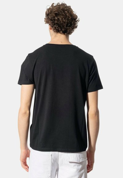 Giovane con capelli ricci che indossa una maglietta nera a maniche corte e pantaloni bianchi, in piedi con la schiena rivolta verso lo spettatore su uno sfondo bianco.