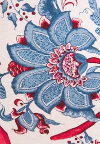 Tissu floral bleu et rouge avec des motifs texturés, présentant des sequins au centre des fleurs et des accents feuillus, sur un fond blanc.