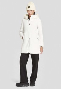 Donna che indossa un cappotto bianco con cappuccio, un berretto in maglia crema, pantaloni neri e sneakers nere, in piedi contro uno sfondo grigio uniforme.