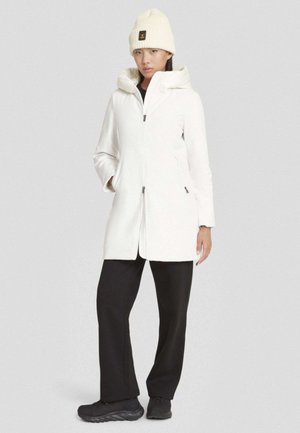 Donna che indossa un cappotto bianco con cappuccio, un berretto in maglia crema, pantaloni neri e sneakers nere, in piedi contro uno sfondo grigio uniforme.