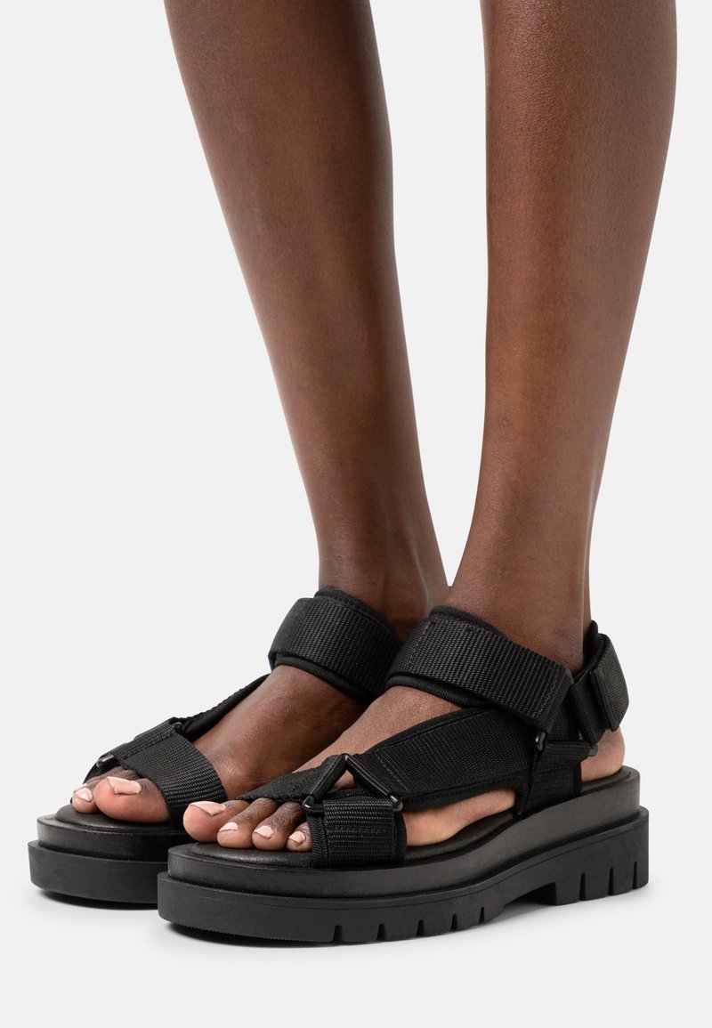 Sandales plateformes noires avec des brides épaisses réglables portées pieds nus avec un teint de peau foncé, montrées de face sur un fond blanc.