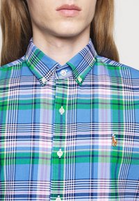 Detailní záběr na osobu, která má na sobě modro-zelenou kostkovanou košili s knoflíky a malým vyšitým logem polo hráče na hrudi.