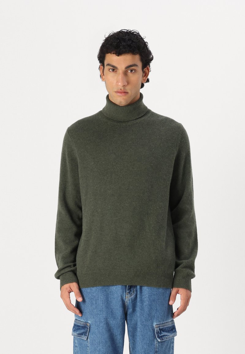 Pull à col roulé vert foncé en matériau tricoté doux, à coupe décontractée, avec des poignets et un ourlet côtelés. Assorti à un pantalon en denim cargo.