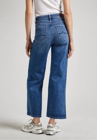 Korkeavyötäröiset leveälahkeiset siniset farkut, joissa on takataskut ja hienovaraisia ompelutietoja. Yhdistetty vaaleanvärisiin lenkkikengiin.