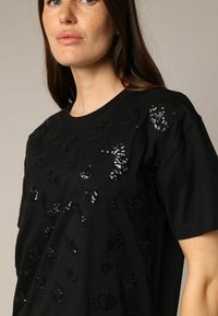 Black t-shirt featuring a short sleeve design, embellished with shiny black sequins in a leopard pattern across the front.