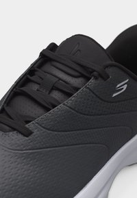 Κοντινό πλάνο ενός μαύρου αθλητικού παπουτσιού με υφασμένο επάνω μέρος, μαύρα κορδόνια, επενδυμένη γλώσσα και λευκή σόλα, με ασημί λογότυπο "S" στο πλάι.