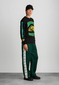 Fekete hosszú ujjú póló zöld és sárga grafikával. Zöld velúr nadrág oldalsó csíkkal, amely logómintákat tartalmaz. Fekete sapka és cipő.