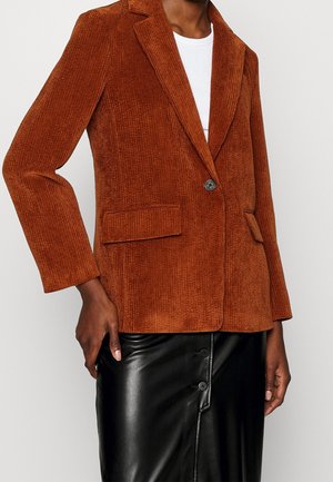 Blazer en velours côtelé couleur rouille avec deux poches à rabat devant, porté sur un haut blanc, associé à une jupe en cuir noire boutonnée.