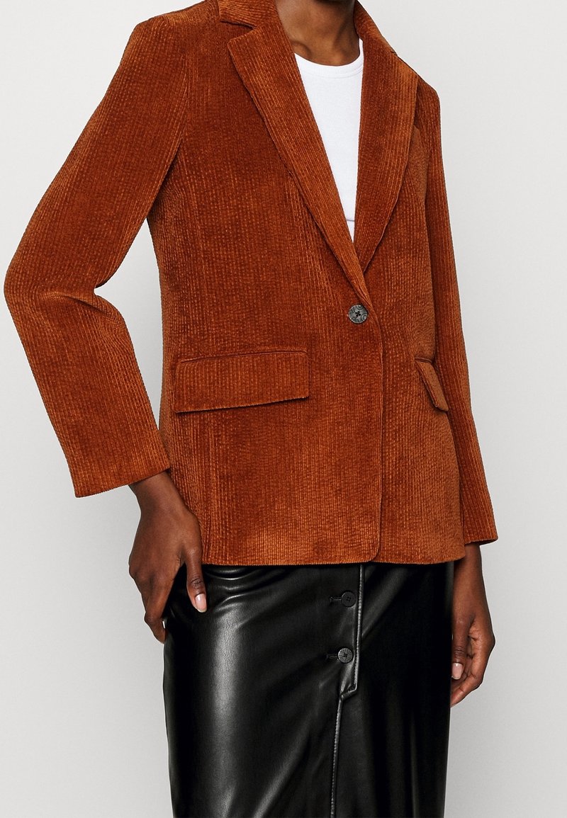 Blazer en velours côtelé couleur rouille avec deux poches à rabat devant, porté sur un haut blanc, associé à une jupe en cuir noire boutonnée.