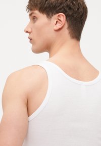 Bijela pamučna majica bez rukava s klasičnim dizajnom, koja ima široke otvore za ruke i okrugli izrez, ističući glatku teksturu i čiste linije.