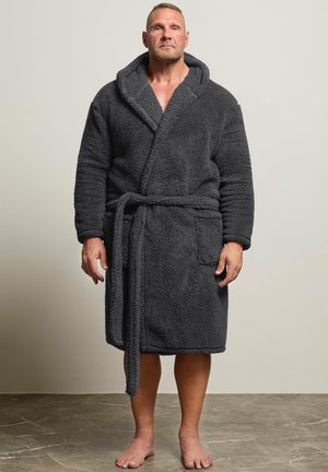Homme debout pieds nus sur un sol, portant un peignoir épais gris foncé avec une capuche et une ceinture nouée.