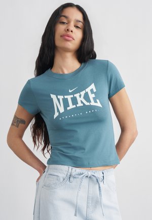 Femeie cu păr lung și închis la culoare, purtând un tricou Nike Athletic Dept. verde-țișo și pantaloni albăstrui deschis cu șnur, pozând pe un fundal deschis.