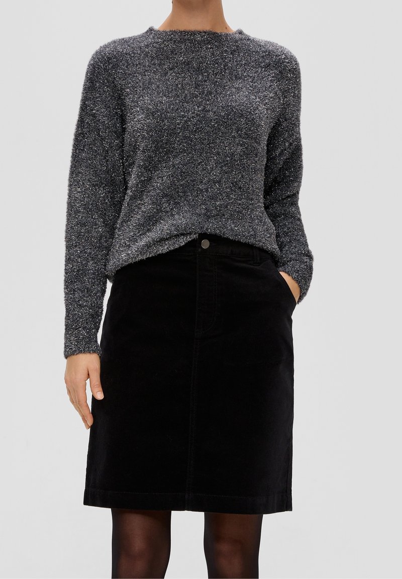 Femme portant un pull long gris en laine douce, rentré dans une jupe noire arrivant au genou, avec une main dans la poche, sur un fond uni.