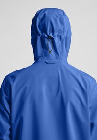 Veste imperméable bleue avec capuche, dotée d'un réglage par cordon, texture lisse et design minimaliste. Vue arrière seulement.