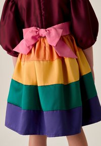 Mehrfarbiges Kleid mit einem lila, grünen, gelben, orangefarbenen und pinken Stufen-Design, einem pinken Satinbogen in der Taille und Puffärmeln.