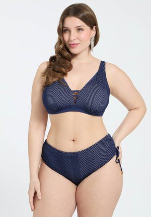 Kobieta z falowanymi brązowymi włosami, ubrana w granatowy, wzorzysty bikini z topem na ramiączkach i majtkami z wysokim stanem wiązanymi po boku, pozująca z ręką na biodrze.