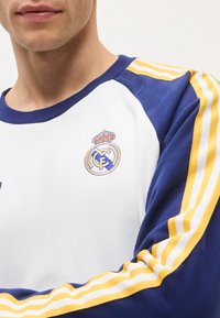 Άνδρας που φοράει λευκό και ναυτικό μπλε μακρυμάνικο πουκάμισο με κίτρινες και άσπρες ρίγες στα μανίκια και λογότυπο της ποδοσφαιρικής ομάδας Ρεάλ Μαδρίτης στο στήθος.