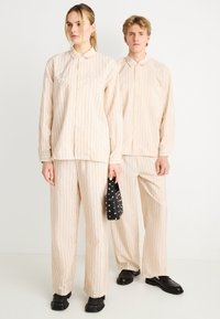 Σετ πουκάμισο και παντελόνι σε μπεζ ριγέ με ελαφρύ ύφασμα. Το πουκάμισο διαθέτει κλείσιμο με κουμπιά και τσέπη· το παντελόνι είναι φαρδύ με ευθύ κόψιμο.