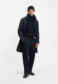 Donkerblauwe trenchcoat met hoge kraag, gebreide coltrui en bijpassende cargobroek. Kenmerken zijn textuur breiwerk, knoopdetails en sneakers.