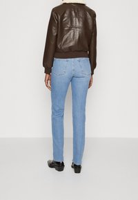 Giacca bomber in pelle marrone con colletto in shearling, abbinata a jeans aderenti blu chiaro e stivali neri con tacco, vista da dietro.