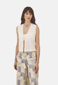 Chaleco de ganchillo blanco con escote en forma de V, bordes decorativos con volantes, cierre frontal con toggle, usado sobre una blusa crema y pantalones amarillos y azules de estampado.