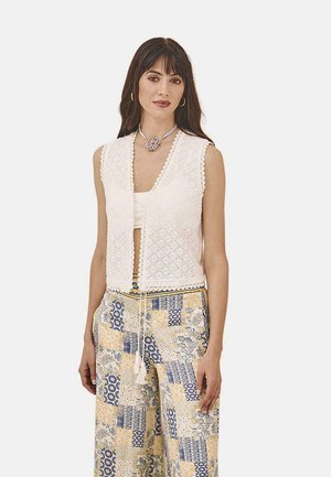 Chaleco de ganchillo blanco con escote en forma de V, bordes decorativos con volantes, cierre frontal con toggle, usado sobre una blusa crema y pantalones amarillos y azules de estampado.