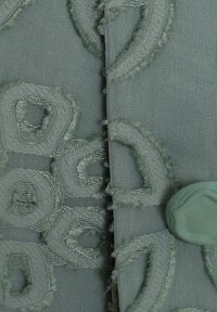 Texturerad ljusgrön tyg med broderade geometriska mönster och en matchande knapp. Mjuka, något fransiga kanter förstärker designen.