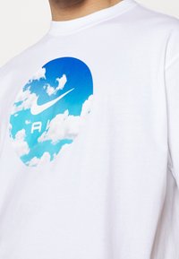 Bílý bavlněný tričko s kulatým modrým grafickým motivem mraků a logem Nike swoosh, s textem "AK" pod ním v kontrastní barvě.