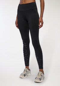 Czarne legginsy sportowe z szerokim pasem w talii, z teksturowanym czarnym wzorem kamuflażu na dolnej części nóg, połączone z szarymi butami sportowymi.