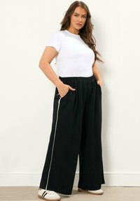 Svarta vida byxor med vita sidostråk, elastiskt midjeband och fickor, tillsammans med en enkel vit t-shirt med kort ärm och avslappnade sneakers.