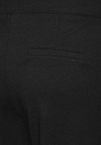 Pantalon noir avec une texture lisse, doté d'une poche passepoilée à l'arrière, de coutures surpiquées et d'une coupe ajustée.