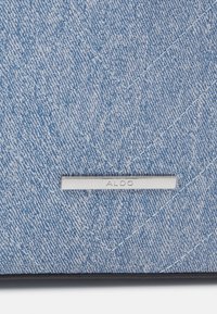 Niebieska powierzchnia o wzorze denimu z ukośnym przeszyciem, z srebrną plakietką z logo ALDO. Teksturowana tkanina, zorganizowany design, czarna lamówka.
