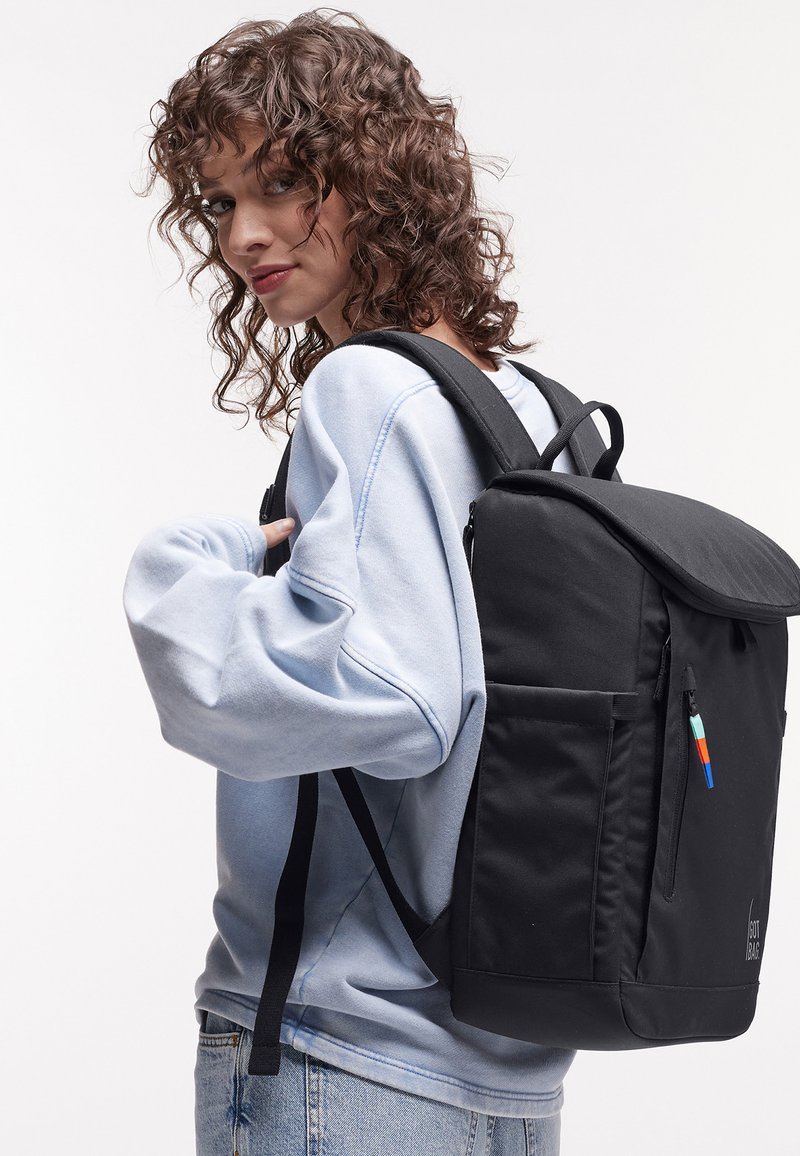 Giovane con capelli ricci che indossa una felpa azzurro chiaro e jeans, porta uno zaino nero sulla schiena su uno sfondo uniforme.
