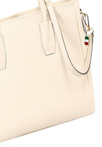 Crèmekleurige leren draagtas met een gestructureerde vorm, subtiele textuur en dubbele handgrepen. Voorzien van een kleine kleurrijke bedel en contrasterende stiksels.