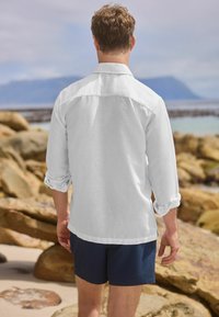 White, long-sleeve shirt with rolled cuffs, paired with navy shorts. Fabric appears lightweight and casual. Background features rocky shoreline.